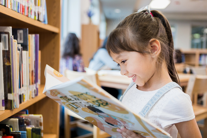 Adorable Hispanic schoolgirl smiles while delight as she reads a picture book while standing in the school library. for article on science of reading